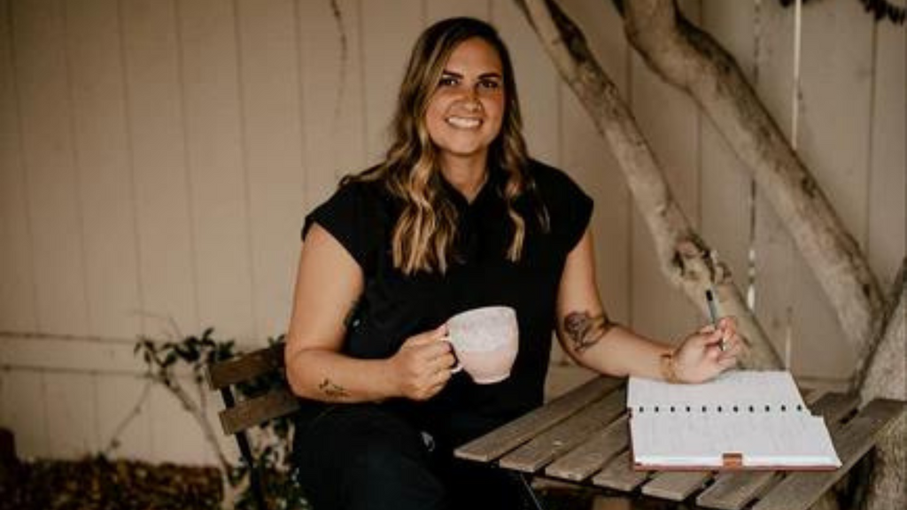 A portrait of Christine Diltz, RN. Christine has long brown hair with dark blonde highlights, and wears navy blue scrubs with light blue BALA Twelves. She is seated at an outdoor coffee table, holding a mug and writing in a notebook.